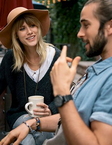 Two friends chat outdoors, woman in a hat smiles while holding a mug, man gestures with his hands as they converse, relaxed vibe.