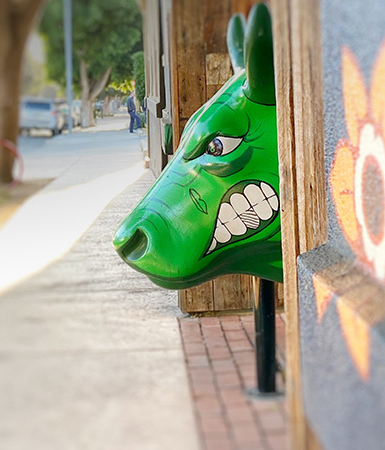 A green dragon mask peeks from a doorway onto a sunny sidewalk, next to a wall mural of a sun.