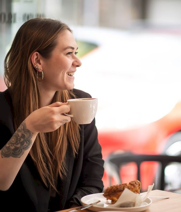 A smiling woman sits at a cafe table, sipping coffee and enjoying a pastry, with a red car blurred outside the window.