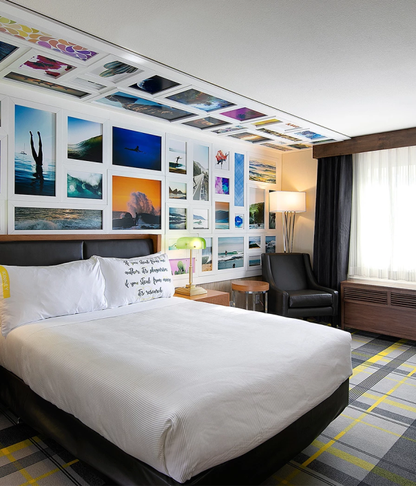 A stylish hotel room with a gallery wall of photos, a comfy bed, lamps, a desk, and a window with curtains.
