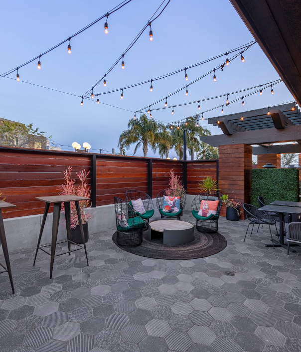 A stylish rooftop patio with string lights, modern seating, a circular rug area, high-top tables, potted plants, and a cozy lounge corner under a covered wall.