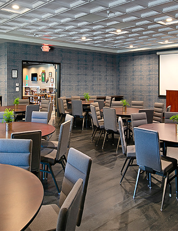 A conference or banquet room with round and rectangular tables, blue chairs, small green plants, a projector screen, and a doorway at the back.