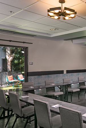 A modern conference room with U-shaped tables and chairs, a large screen at the front, ceiling lights, and large windows showing an outdoor area.