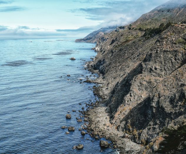 Cliffside rocks meet calm blue sea along a rocky coastline, with clear skies above.