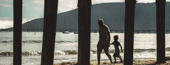 Two people walking on the beach at sunset under a pier, framed by columns.