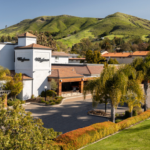 A hotel and clubhouse complex with white buildings, palm trees, manicured lawns, and a scenic green hillside in the background.