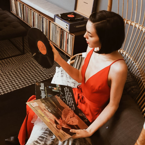 A woman in a red dress sits on a couch, holding a vinyl record and a magazine, surrounded by a retro setup with shelves and a turntable.
