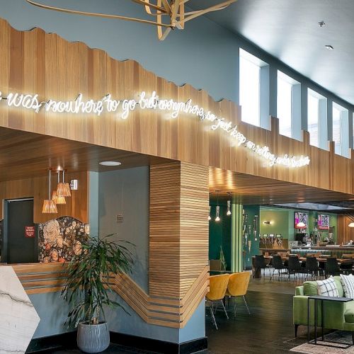 A stylish, sunlit hotel or cafe lobby with wood paneling, a neon quote along the ceiling, comfy seating, plants, and a bookshelf area.