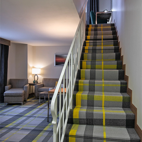 A hotel lobby with a carpeted staircase featuring yellow and gray stripes, a seating area with chairs and a lamp, and soft wall lighting.