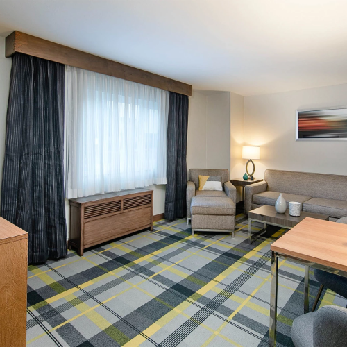 A hotel room with a wooden desk, large windows with dark curtains, a beige sofa, armchairs, a coffee table, a lamp, and a plaid carpet; cozy lighting.