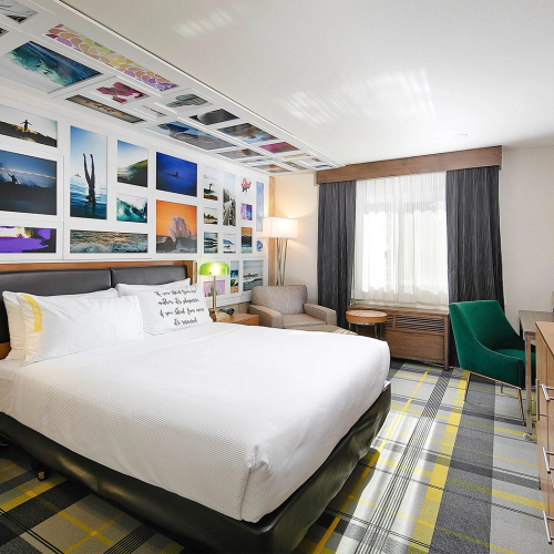 Two lamps, a king bed, and a gallery wall of colorful photos above the headboard, plus a desk, TV, and a bright seating area in a modern hotel room.