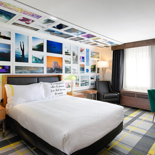 A modern hotel room with a gallery wall of colorful photos, a comfy bed, two nightstands, a desk, and a plaid carpet, ending with a cozy vibe.