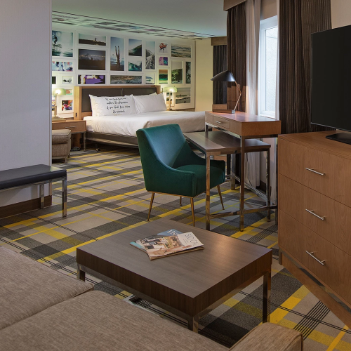 A modern hotel suite with a bed, sofa, green armchair, coffee table, wooden dresser with a large TV, and a collage wall above the bed.