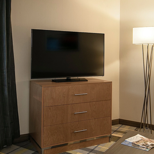 A living room corner with a wooden dresser supporting a TV, a tall floor lamp, and a glass-top table; neutral tones and a magazine on the coffee table.