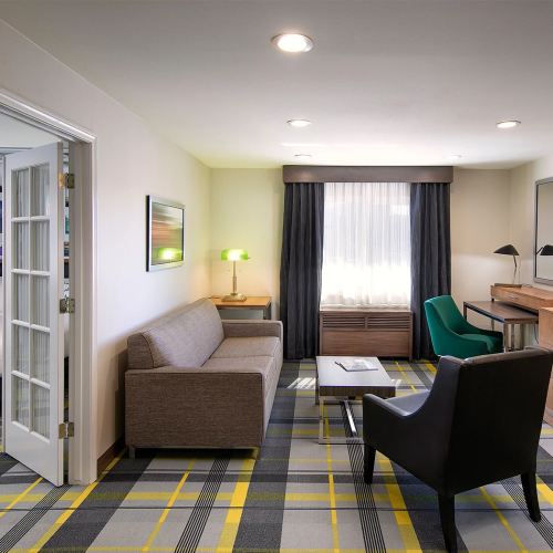 A modern hotel suite with a living area, sofa, armchairs, coffee table, TV on a wooden dresser, and a window with dark curtains; a connected bedroom is visible through a doorway.