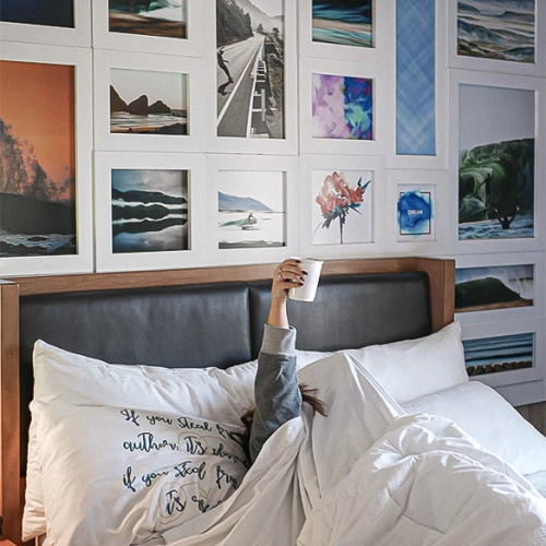 Person in bed under white duvet, head unseen, holding up a phone; wall art collage of ocean, landscapes, and abstract prints behind.
