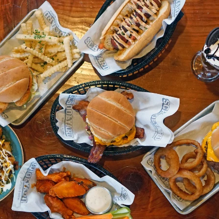 A spread of burgers, fries, onion rings, salad, dipping sauces, and a milkshake on a wooden table, seen from above.
