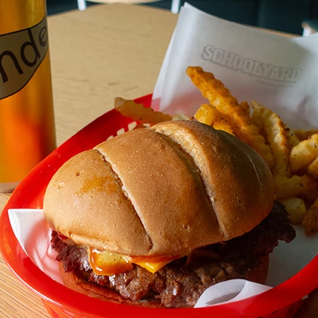 A cheeseburger with onions on a bun, crispy fries in a red basket, and a bottle of Blonde beer on a wooden table.