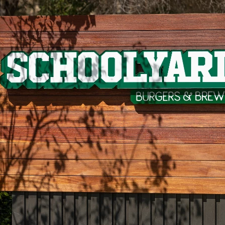 Schoolyard sign advertising burgers & brew, mounted on a wooden panel with sunlit shadows.