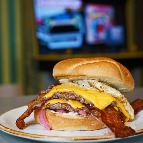 A loaded bacon cheeseburger with onion rings, melted cheese, and condiments on a bun, on a plate near a game arcade backdrop.