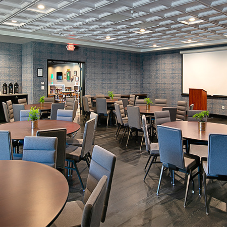 A spacious conference room set with round and square tables, gray chairs, small potted plants, a projector screen, and a coffee bar near the back, ready for events.