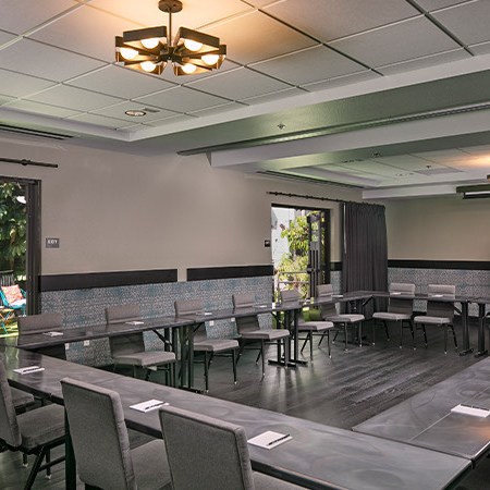 A modern meeting room with U-shaped and long tables, grey chairs, a large screen, and a doorway to a outdoor area; soft lighting and tidy setup.