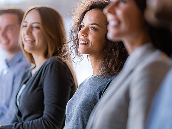 A group of diverse people seated in a row, smiling and looking to the side, likely at a presentation or event, engaging and attentive.