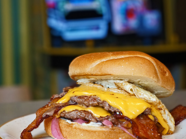A towering bacon cheeseburger with two beef patties, melted cheddar, onions, and crispy bacon, on a bun, served on a plate.